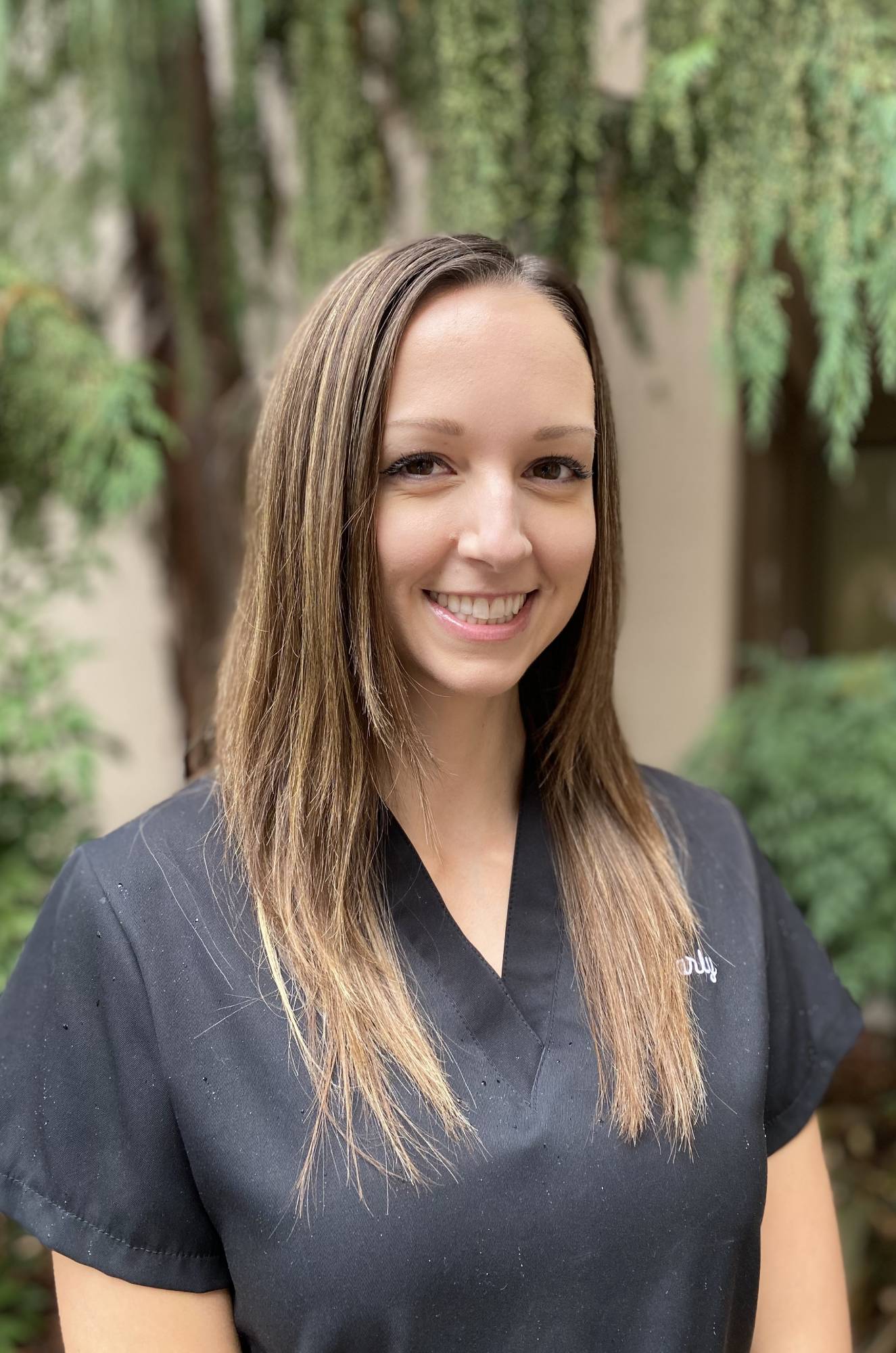 Smiling woman in dark shirt poses outdoors with green foliage background for orthodontics practice.