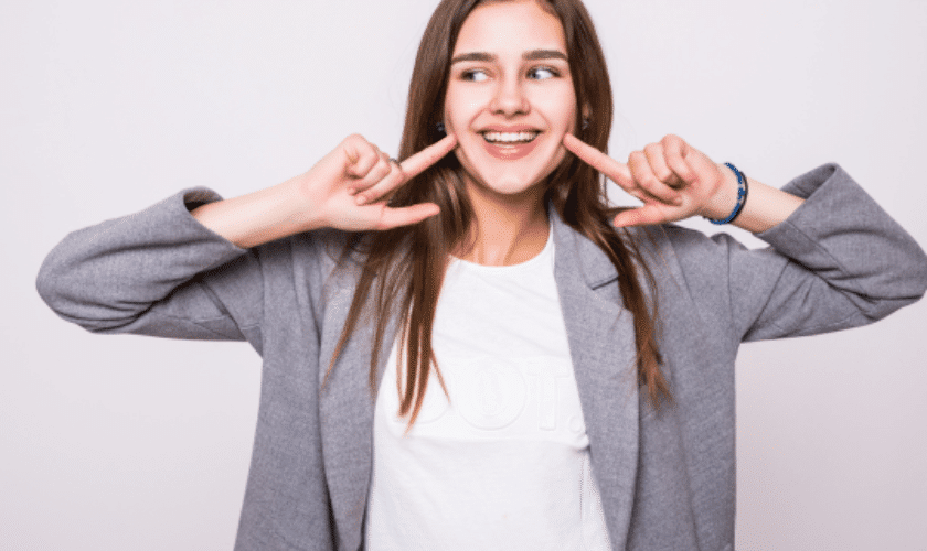 Woman in business casual attire smiling while pointing at her teeth against white background.