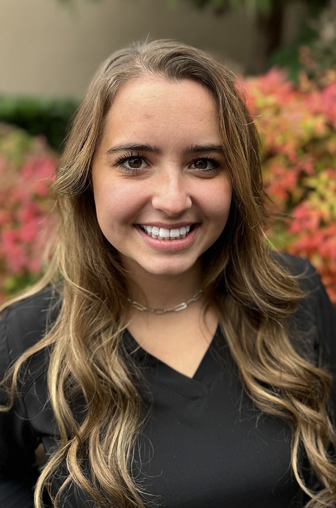 Young woman smiling at camera with flowers in background, professional portrait for orthodontics practice.