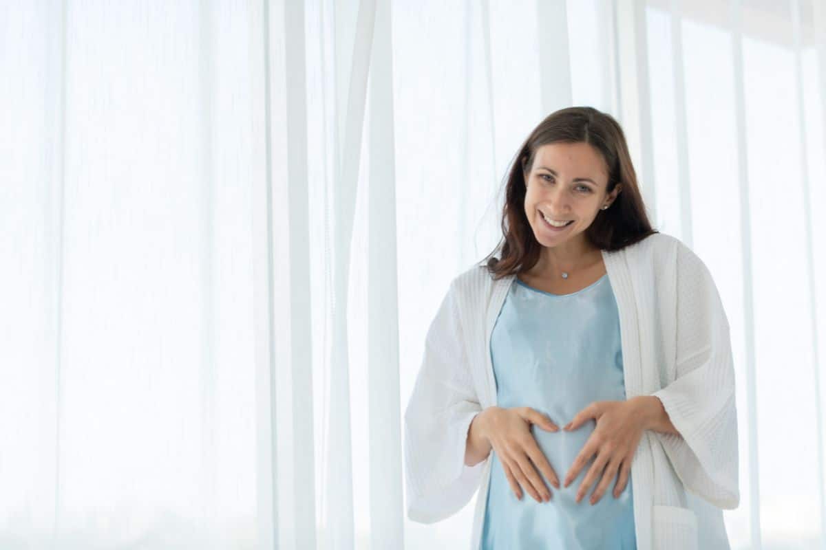 Woman in blue top and white cardigan standing in bright, windowed room touching her stomach.